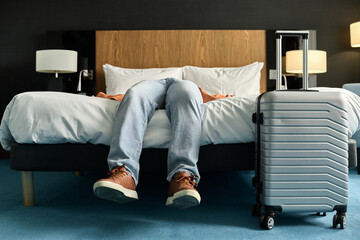 Low section of unrecognizable man lying on bed in hotel room exhausted after travelling focus on feet wearing leather sneakers and suitcase copy space - Powered by Adobe
