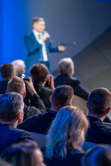 Speaker is addressing an audience at a business conference. People in the audience are paying attention and some are taking photos.