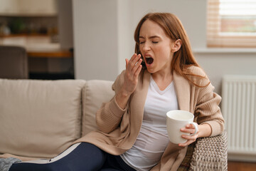 redhead woman yawning drinking coffee tea sitting on sofa at home