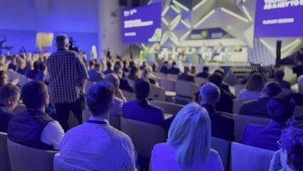 Modern conference hall filled with attendees focused on a speaker. The setting features futuristic designs and professional lighting, illustrating an engaging business event.