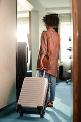 Vertical back view of female guest entering hotel room and rolling suitcase arriving at resort