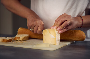 Close-up hands of a woman slicing cheese on cutting board, using a kitchen knife, preparing...