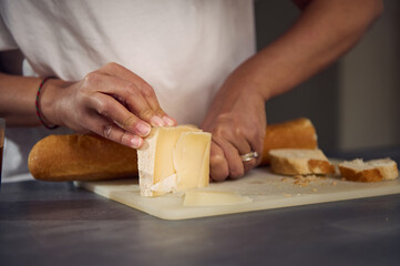 Close-up hands of a woman slicing cheese on cutting board, using a kitchen knife, preparing...