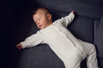 View from above of a cute baby boy in white bodysuit, sweetly sleeping on comfortable gray sofa