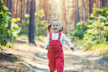 Fototapeta premium Portrait of smiling three years old caucasian child girl runs on the trail in the pine forest. Waving hands. Forest on bright light background.