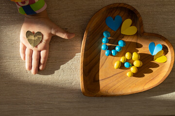 On child s hand lies heart cut out of military camouflage fabric and yellow and blue letters UA, lined with candies. Stop war in Ukraine. With love and gratitude to the soldiers of the Armed Forces