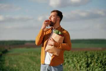 Eating an apple, holding shopping bag. Handsome man is on the agricultural field at daytime