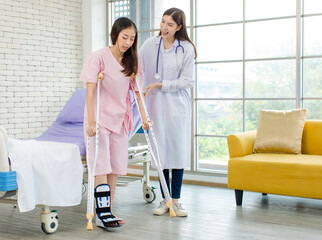 Asian female patient with broken leg assisted by woman doctor. Injured young woman stands up from...