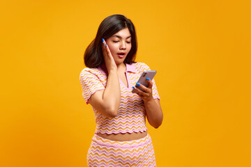 Young Indian woman holding smartphone and looking to its screen with shocked expression against...