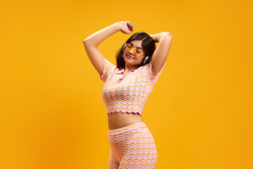 Active brunette woman dancing raising hands while listening music in headphones against vibrant yellow studio background. Concept of human emotions, fashion and style, music and dance, youth, beauty.