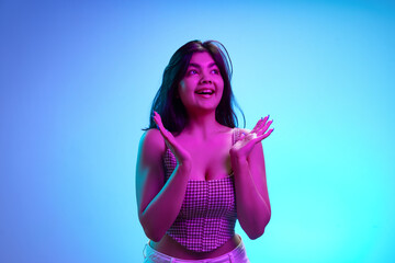 Young brunette Indian woman clapping hands of joy, excitement in pink neon light against gradient blue background. Concept of human emotions, beauty and fashion, youth. Ad