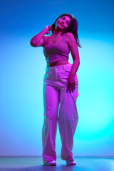 Young brunette Indian girl dancing of joy in motion in pink neon light against gradient blue...