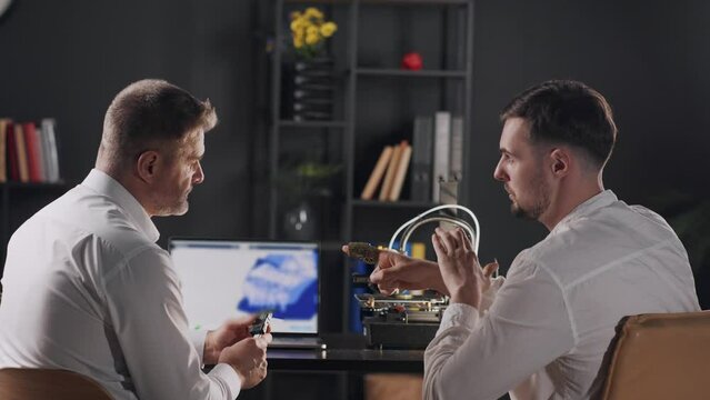 Two medical engineers uses digital calipers and measures the parameters of 3d printed prosthesis. A middle aged man graphic designer discuss printed work. A 3d printer makes prosthesis for human