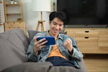 Happy asian man sitting on couch and celebrating victory in mobile game with raised fist