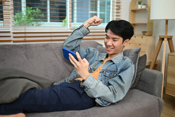 Young man relaxing on couch at home and looking at cellphone celebrating online prize win