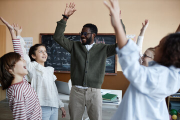 Young African American teacher and group of kids raising arms while having fun during lesson in...