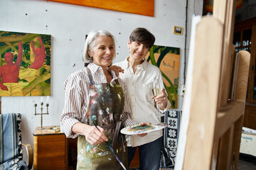 A mature lesbian couple stands in an art studio, sharing a moment of togetherness.