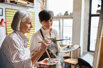 Two women immersed in creativity, paint with a brush and palette in an art studio.