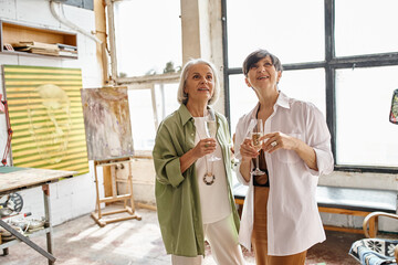 Two mature women, a loving couple, stand side by side in an art studio.
