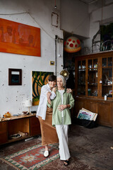 Two mature women stand in a room with a painting on the wall.