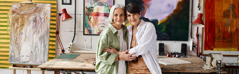 Two mature women, a couple, standing stylishly together in an art studio.