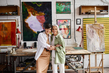 Two mature women, a lesbian couple, stand side by side in an art studio.