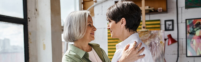 A mature lesbian couple standing confidently side by side in an art studio.