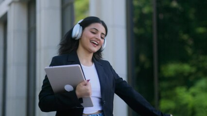 Charming young indian businesswoman with headphones listening to music while walking down the street after work outdoors Happy relaxed lady on the city centre enjoying beautiful day alone