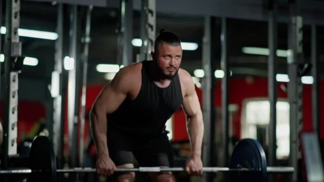 Muscular man in black tank top performing bent-over row with barbell, demonstrating strength and determination in gym