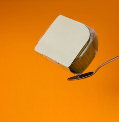 Yogurt cup balanced on spoon against orange background