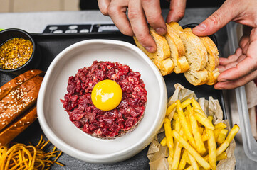 A close-up of a hand placing sliced bread on a plate with raw minced meat topped with an egg yolk, next to fries and condiments