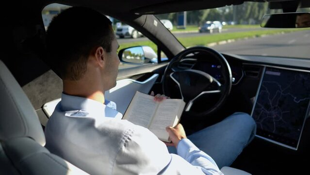 Male businessperson reading book during riding on electrical vehicle with autopilot at urban road. Successful businessman improving his knowledge while riding an autonomous self driving electric car