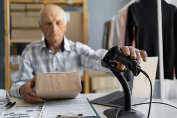 Elderly male shop owner sitting in front of desk, using computer and barcode scanner, recording...