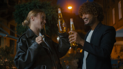 European woman and Arabian Indian Latino man open bottle of beer cider clink glasses walk street outside city evening friends drinking smiling weekend multiracial couple celebrate happy together enjoy
