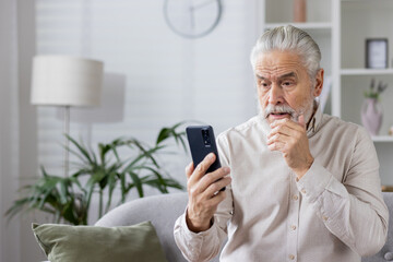 A serious senior male businessman is holding a phone in his hands and looking worriedly at the screen, rubbing his gray beard with his hand, talking on a video call