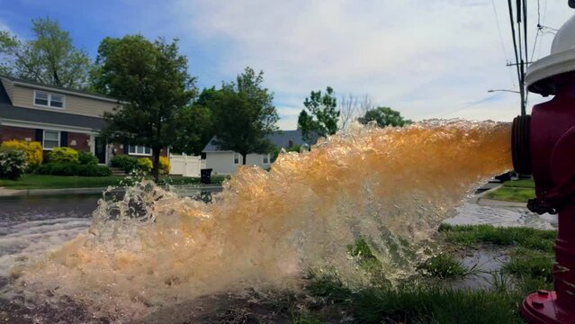 A low angle shot of a red and white fire hydrant spraying rusty water onto the street on a sunny day. Filmed in slow motion, the camera truck left beside the hydrant as it floods the road.