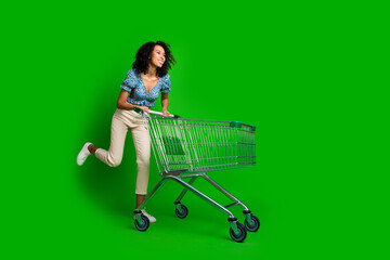 Full length portrait of pretty young girl push trolley empty space wear top isolated on green color...