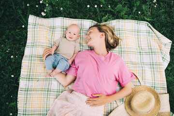 Smiling mom and baby lying on blanket on green grass at summer. Family relaxing and having picnic outdoors. Beautiful mother with her baby on nature. Concept of motherhood, human happiness, eco life.