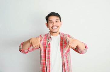 Happy young Asian man in casual shirt pointing finger down and smiling at camera isolated on white background.
