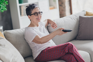 Photo of nice senior woman hold remote controller sit sofa dressed white clothes cozy living room...