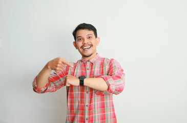 Portrait of excited Asian man in casual shirt looking at his wrist watch, pointing on it to remind the time. Advertising concept. Isolated image on white background