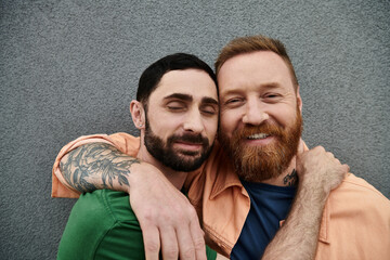 Two men embrace lovingly in front of a grey wall, expressing their deep connection and affection...