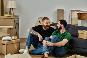 Gay couple sitting on wooden floor, surrounded by moving boxes, starting new life together in cozy...