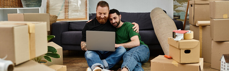 A man and a woman sit on the floor, deeply focused on a laptop screen, amidst unpacked boxes in...