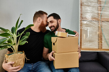 Gay couple in love, sitting on couch surrounded by boxes, holding boxes and plant, moving into new...