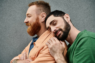 Gay couple in casual attire, two men with beards, hug each other against a grey wall in a display...