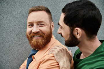 A gay couple in casual attire stand side by side against a grey wall, showing their love for each...