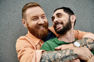 Two men with tattoos on arms, in casual attire, embrace in love, standing against grey wall.