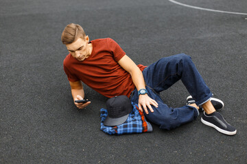 handsome fashionable student guy in stylish casual clothes with jeans lies on the asphalt and uses...