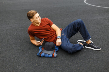 stylish handsome young guy in fashionable clothes with a T-shirt and jeans lies on the asphalt at the stadium with a smartphone